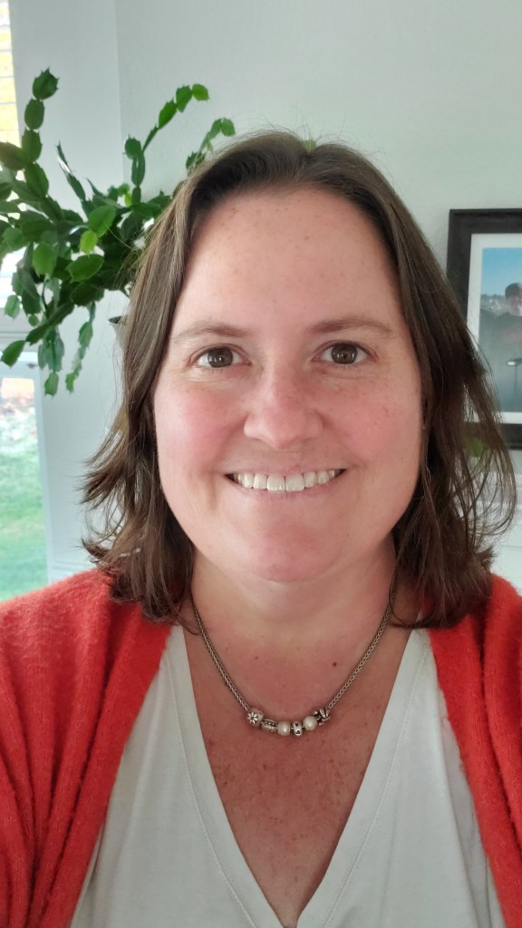 Image of Susan wearing an orange sweater over a white shirt, standing in front of a plant and framed photo.